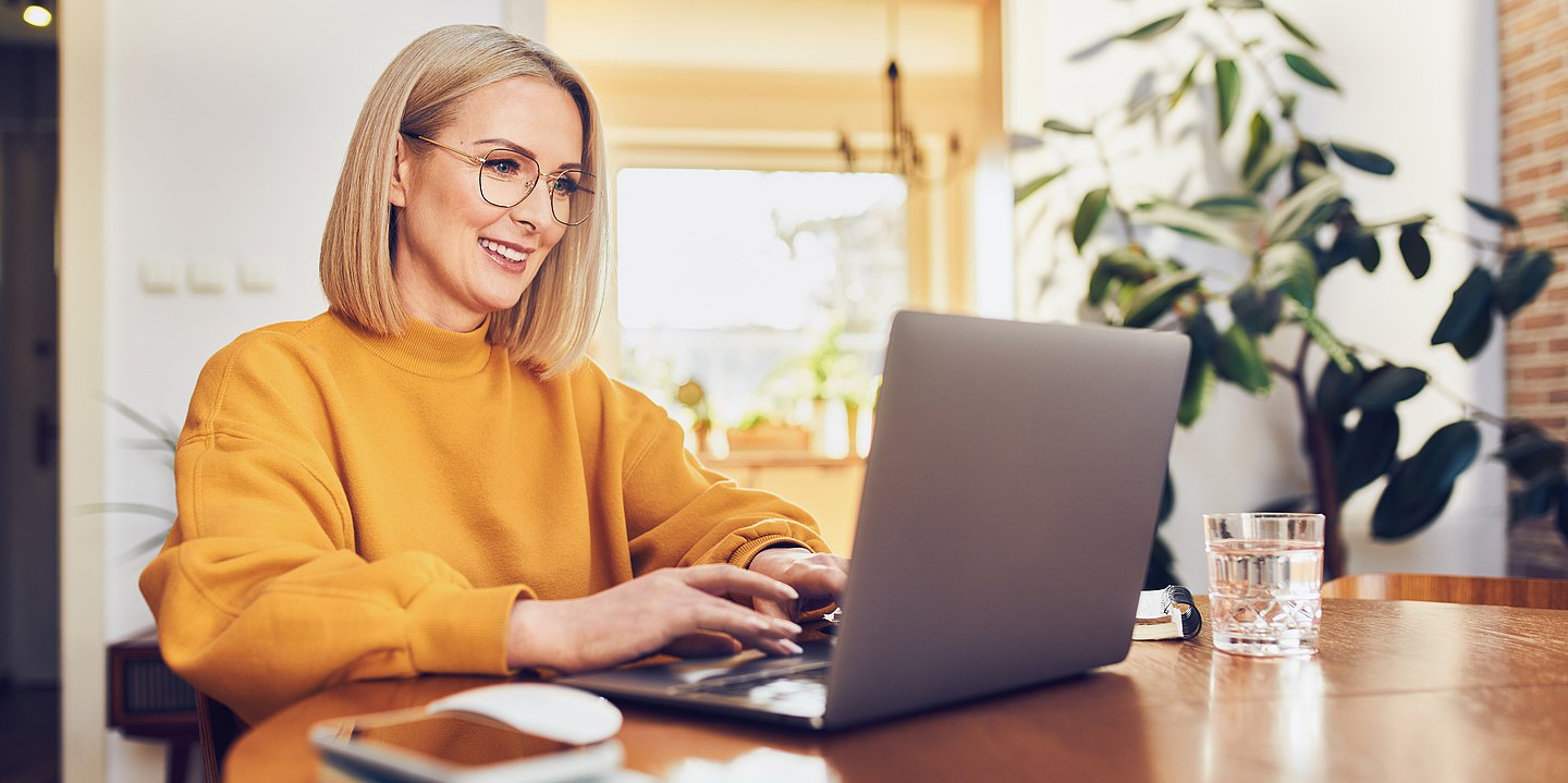 Das Bild zeigt eine blonde Frau mit Brille am Küchentisch vor einem Laptop bei der Recherche. Sie trägt einen dunkelgelben Pullover. Rechts neben dem Laptop steht ein Glas mit Wasser.