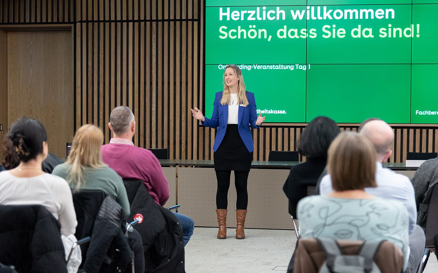 In einem mit Holz vertäfelten Saal sitzen zahlreiche Männer und Frauen und blicken auf eine kleine Bühne. Dort wird eine Begrüßung der Versammelten an die Wand projiziert.