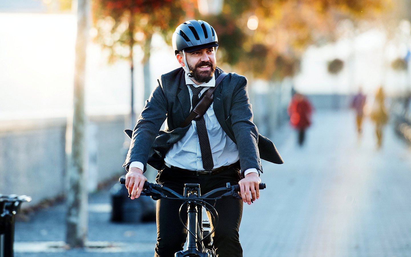 Foto eines radelnden Mannes in Businesskleidung, der einen Weg in der Stadt entlang fährt und auf dem Weg zur Arbeit ist.