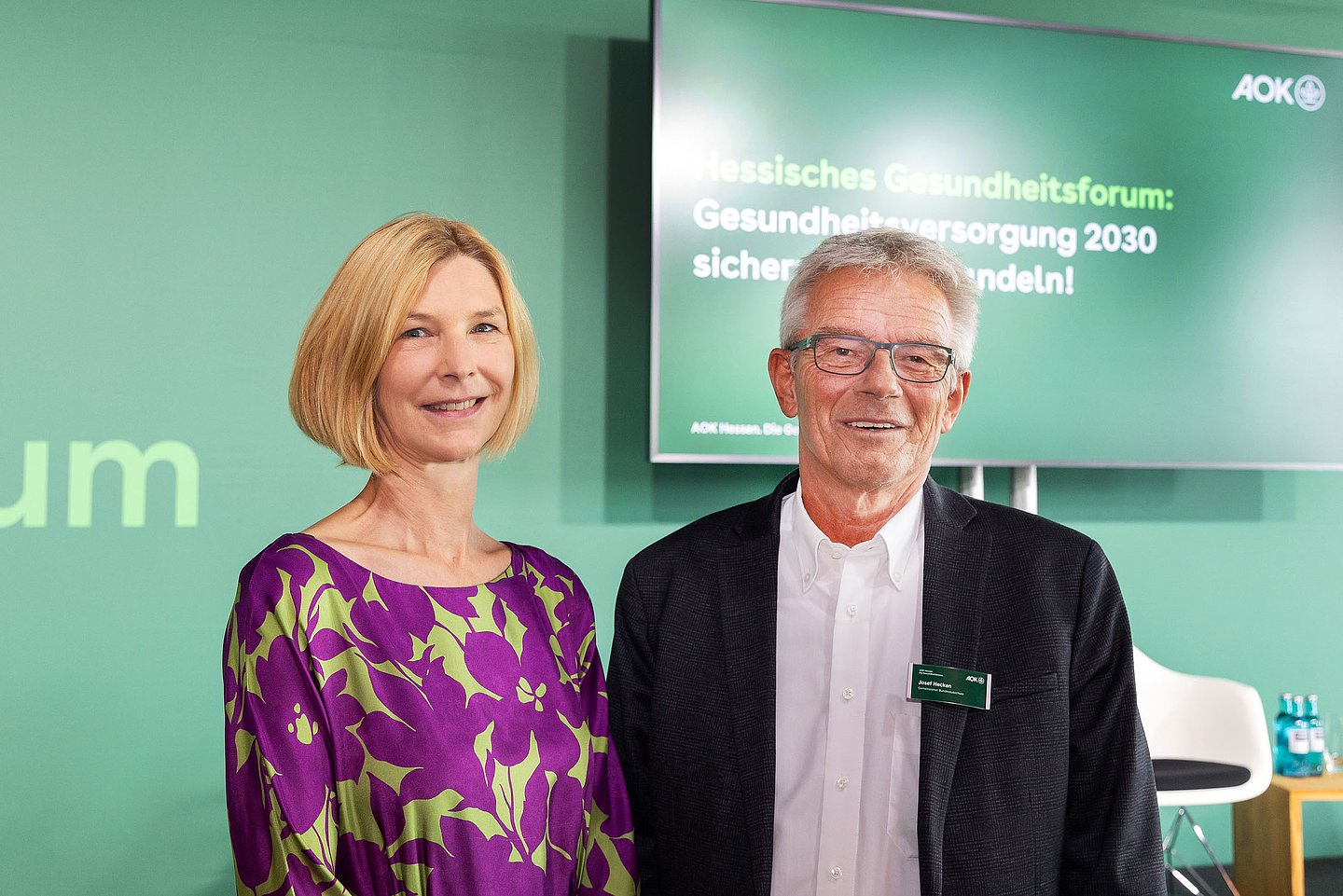 Das Bild zeigt eine blonde Frau (links) in violetter Bluse mit hellgrünen Farbflecken und einen graumelierten Brillenträger mit dunkelgrauem zart karierten Sakko und offenem weißen Hemd.