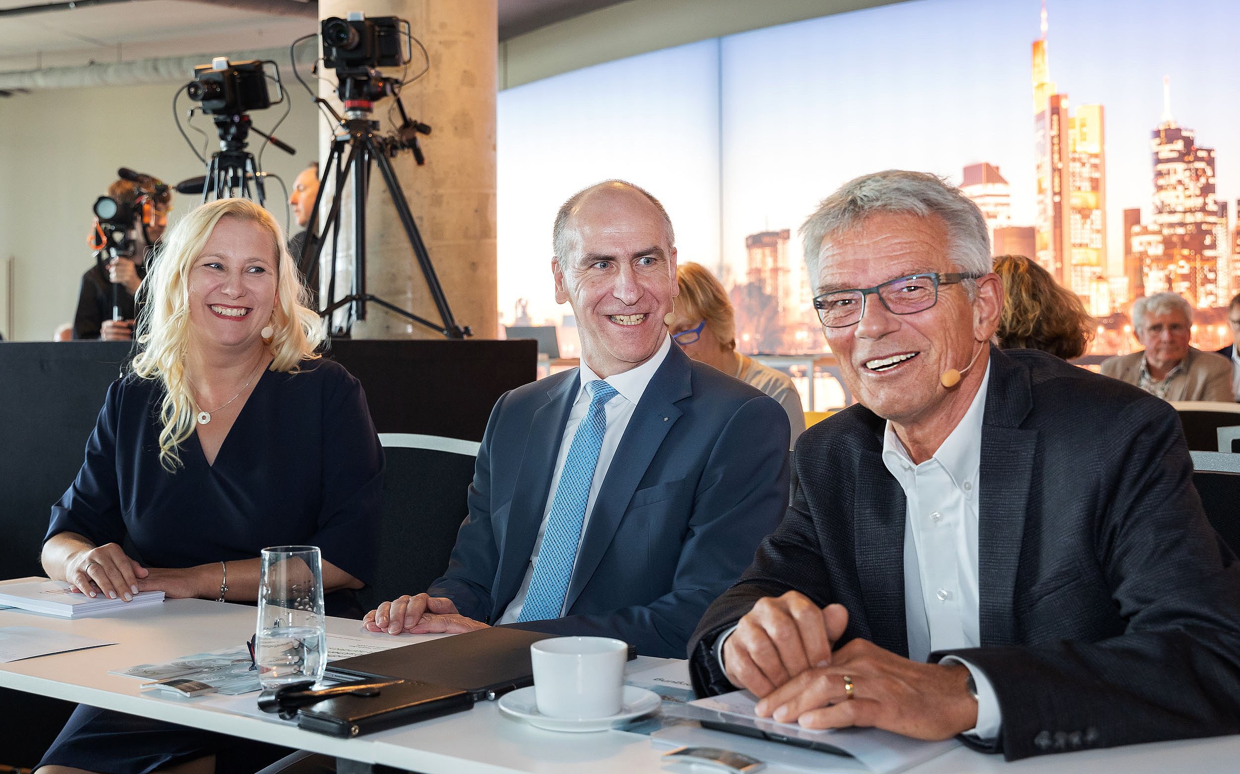 Das Bild zeigt von links nach rechts eine blonde Frau mit langen Haaren und dunkelblauem Kelid, einen Mann mit Halbglatze stahlblauem Sakko, weißem Hemd und hell-dunkelblau-karrierter Krwatte, einen Mann mit vollem grau-melierten Haar, blau-karierten Sakko und offenem weißen Hemd., 