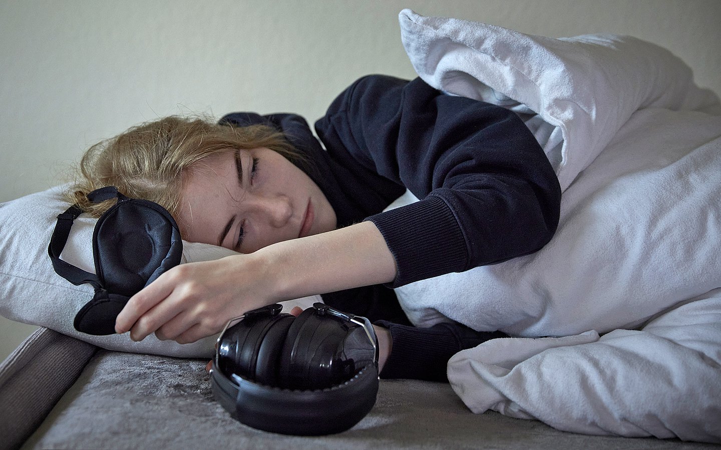 Foto einer jungen Frau, die völlig erschöpft im Bett liegt mit einer Schlafbrille in der einen und Kopfhörern in der anderen Hand