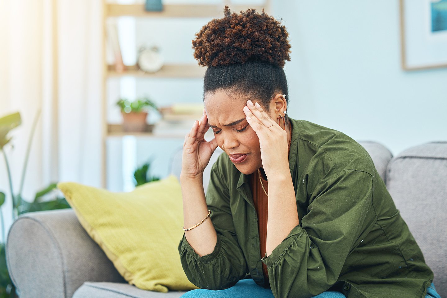 Eine junge Frau sitzt leicht vorgebeugt auf einem Sofa. Die Augen sind geschlossen, das Gesicht wirkt angespannt und sie fasst sich mit beiden Händen an den Schläfen. Die Arme sind dabei auf den Knien abgestützt.