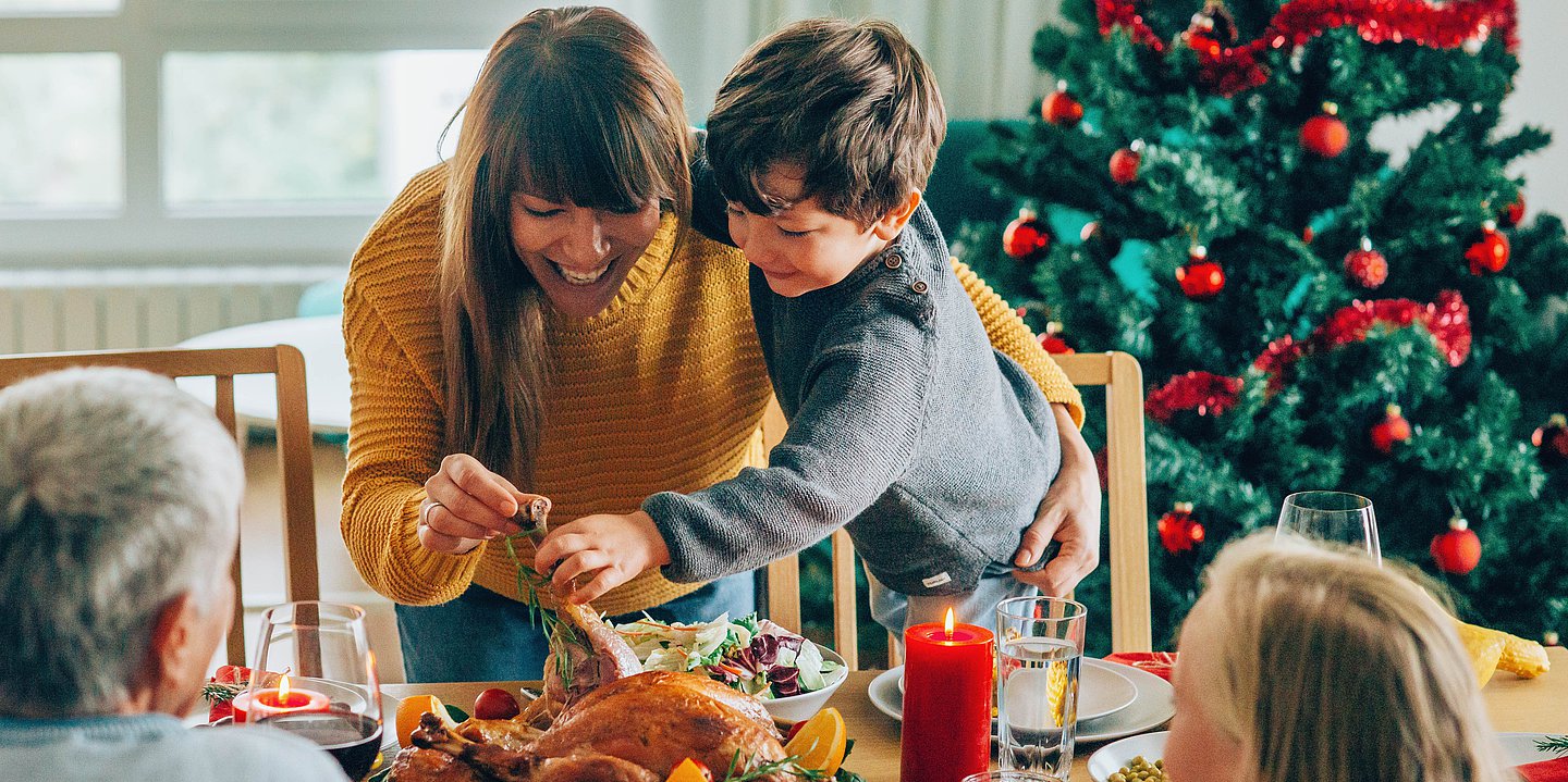 Eine Familie beim Festtagsessen, eine junge Frau und ein Kind verteilen lachend Essen, ein geschmückter Weihachtsbaum steht im Hintergrund.