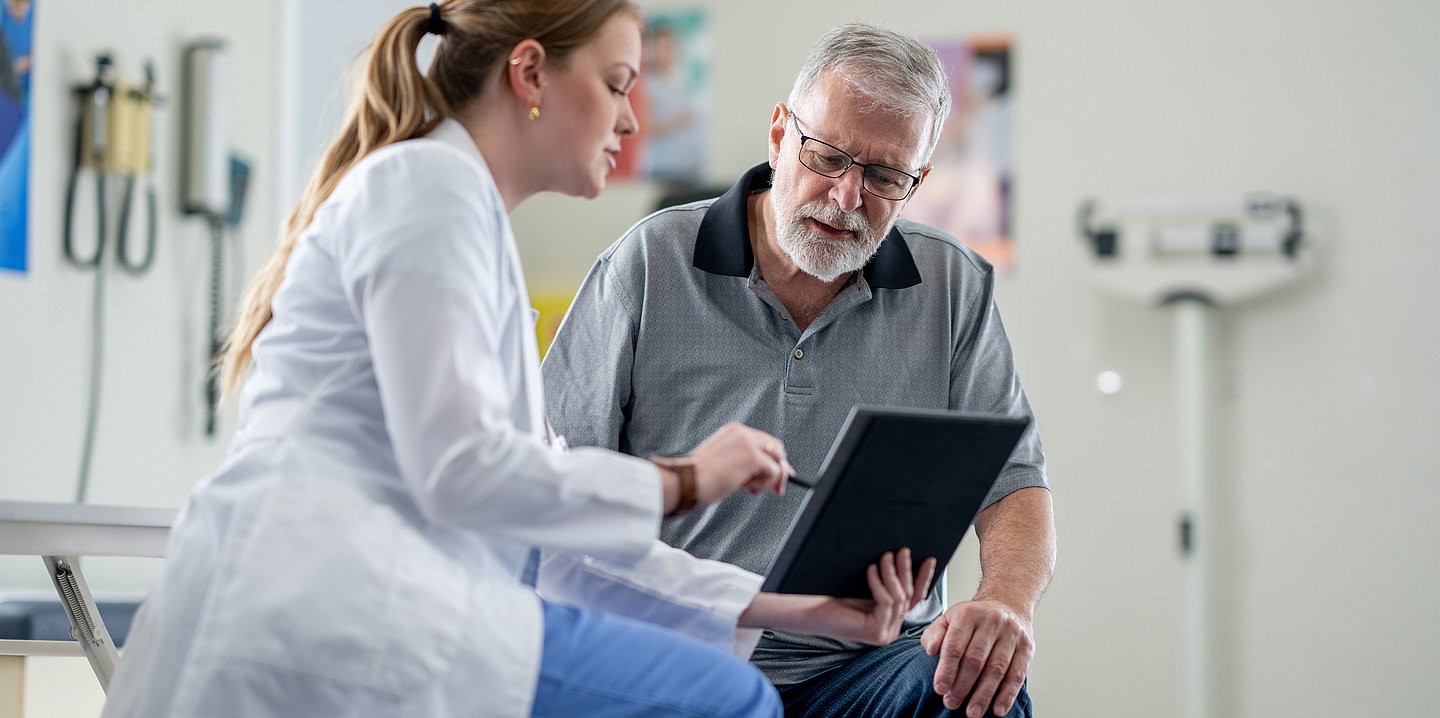 Foto: Eine Ärztin im weißen Kittel hält ein Tablet in der Hand und deutet darauf, daneben sitzt ein Mann im mittleren Alter und schaut ebenfalls auf das Gerät.