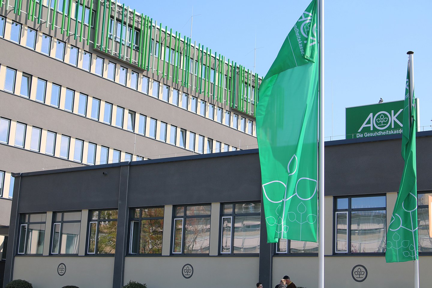 Das Bild zeigt ein großes grau-braunes Verwaltungsgebäude im Anschnitt mit drei parallelen Fensterreihen mit. Oben schließt der Bau mit grünen Längsstreben ab. Vor dem Hauptgebäude steht eine Art Büro-Pavillion, auf dem Dach ein AOK-Schild, davor zwei Masten mit grünen AOK-Fahnen.