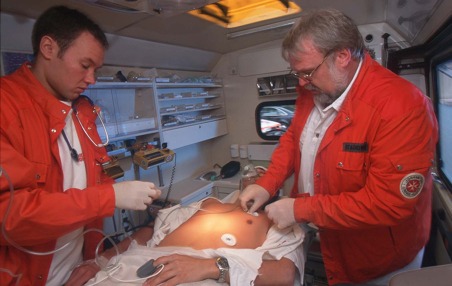 Das Bild zeigt zwei Rettungssanitäter mit orangen Rettungsjacken beim EKG im Rettungswagen. In der Mitte liegt ein Patient mit Sauerstoffmaske, dem die Pads des EKG angelegt werden. Am linken Finger trägt er einen Pulsmesser.