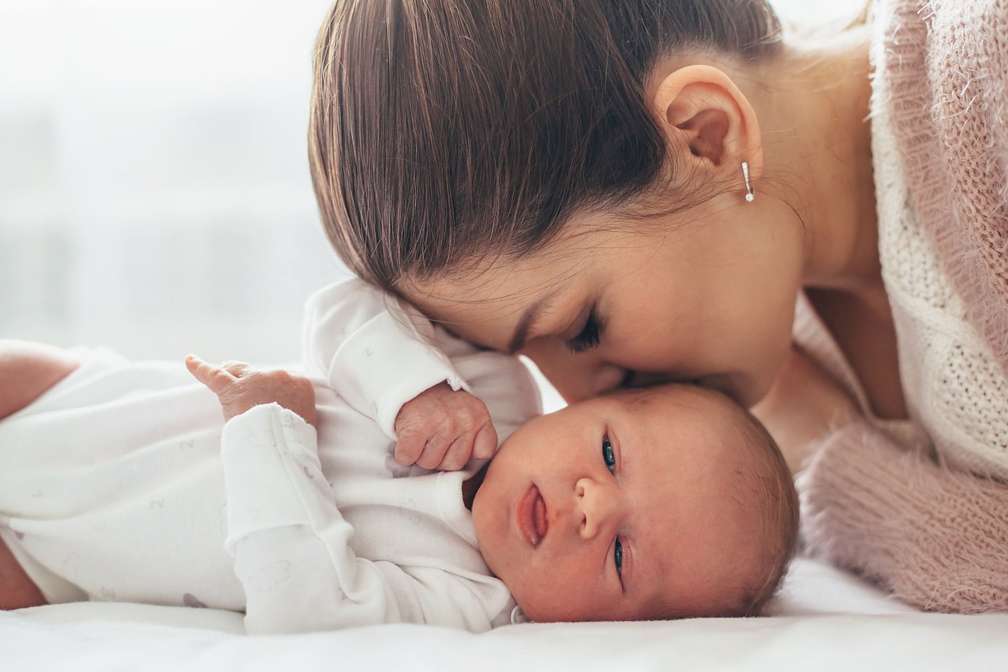 Das Foto zeigt ein Baby auf einer Unterlage, die Mutter beugt sich von der Kopfseite her über ihr Baby und liebkost es seitlich am Kopf.und gt