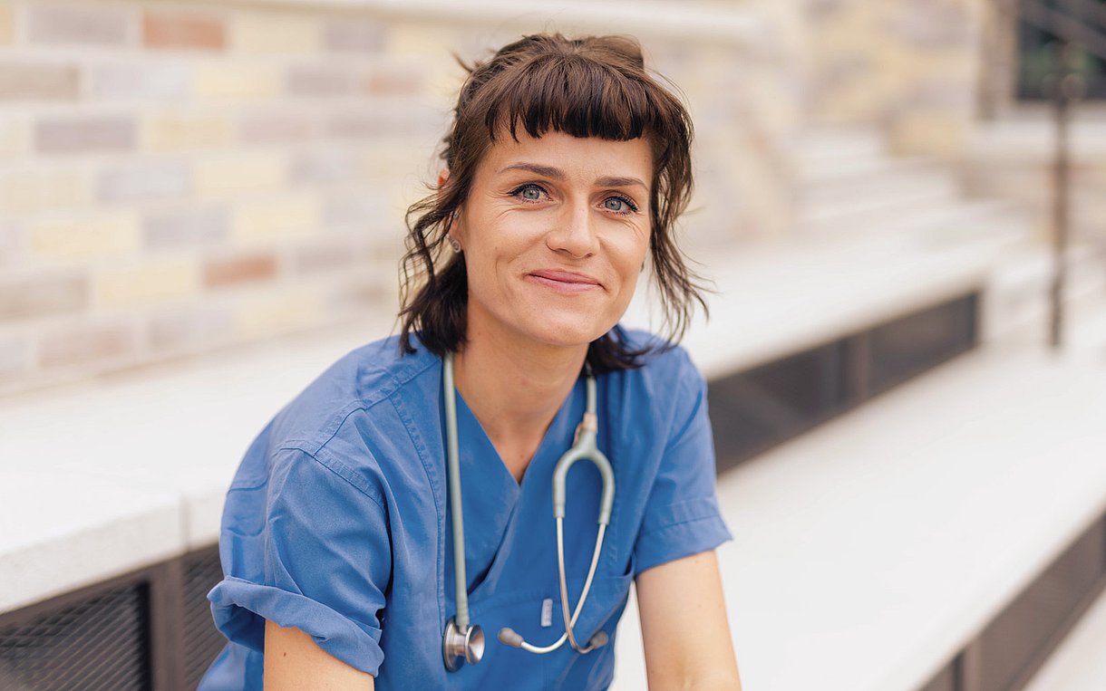 Die Gesundheits- und Krankenpflegekraft, Jenny Kunert sitzt in ihrer Berufskleidung draußen auf der Treppe und schaut in die Kamera