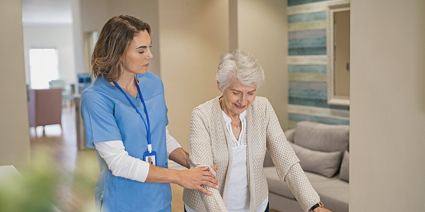 Eine Pflegefachkraft hilft einer älteren Dame, mit einem Rollator, beim Laufen