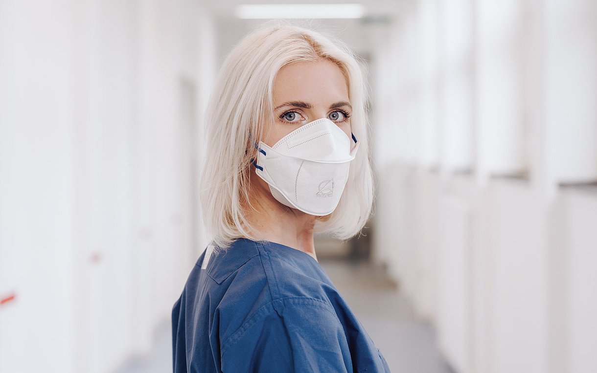 Das Bild zeigt die Gesundheits- und Krankenpflegerin Vanessa Schulte im Klinikflur, zurückschauend in die Kamera mit Maske