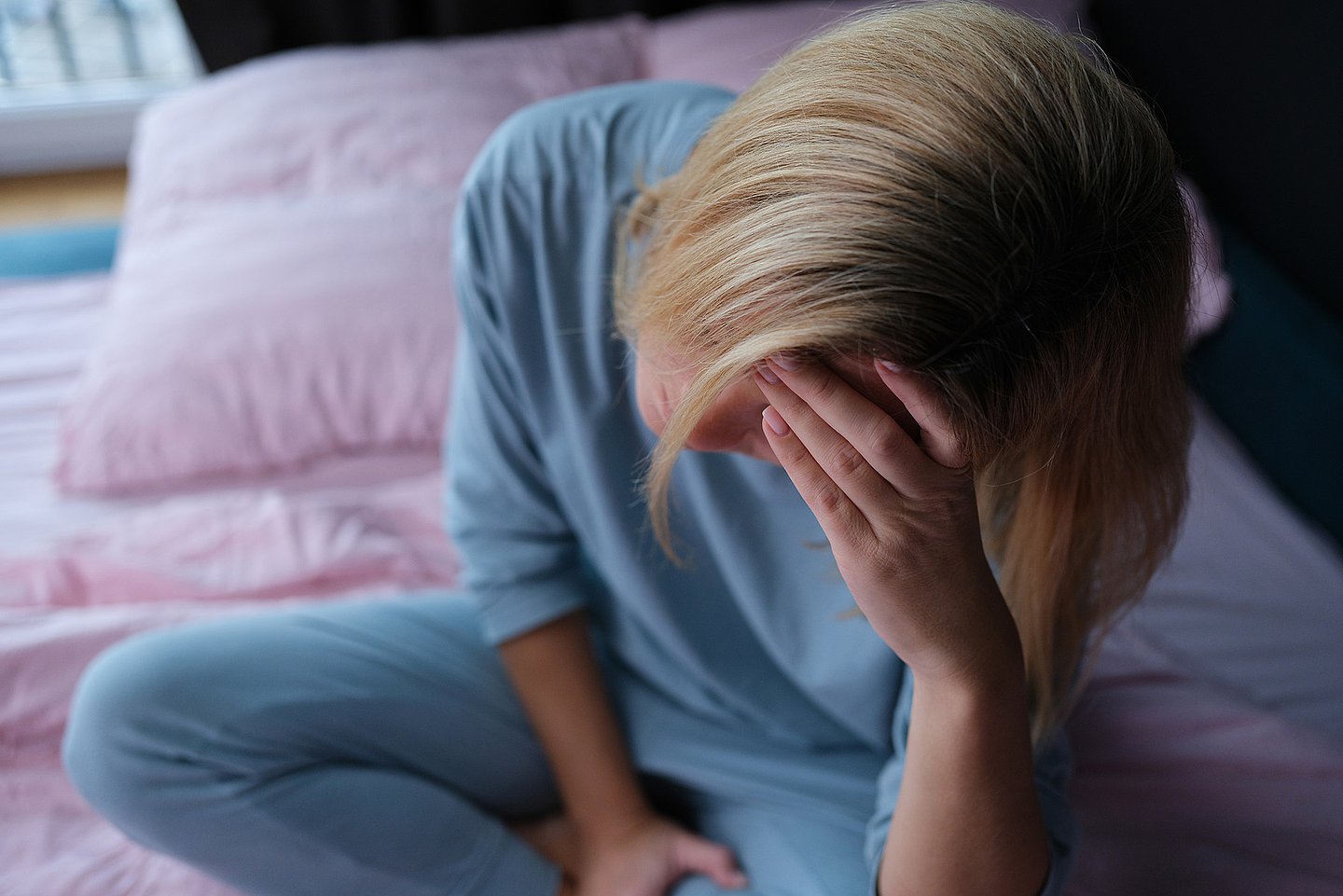 Ein Frau in einem blauen Schlafanzug sitzt mit gekreuzten Beinen auf ihrem Bett, die linke Hand an den Kopf gestützt, den Blick nach unten gerichtet.