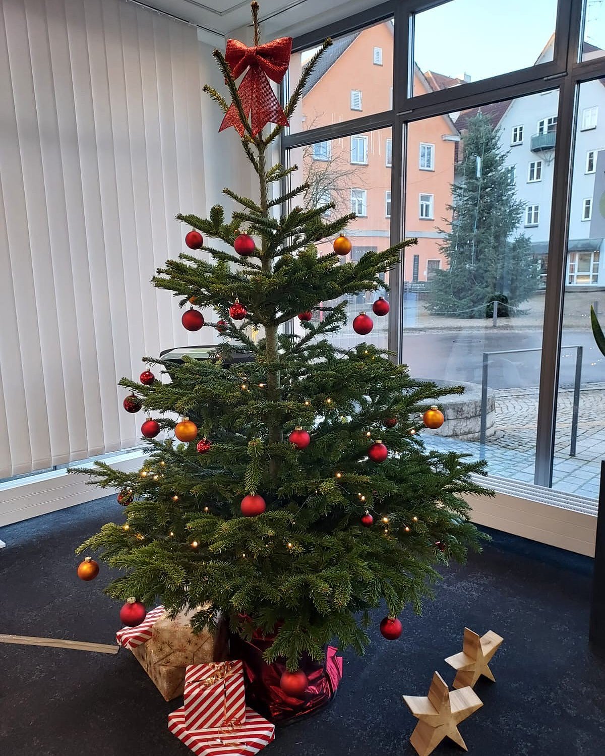 Ein geschmückter Nadelbaum, unter dem Geschenke liegen, steht vor einem Fenster.