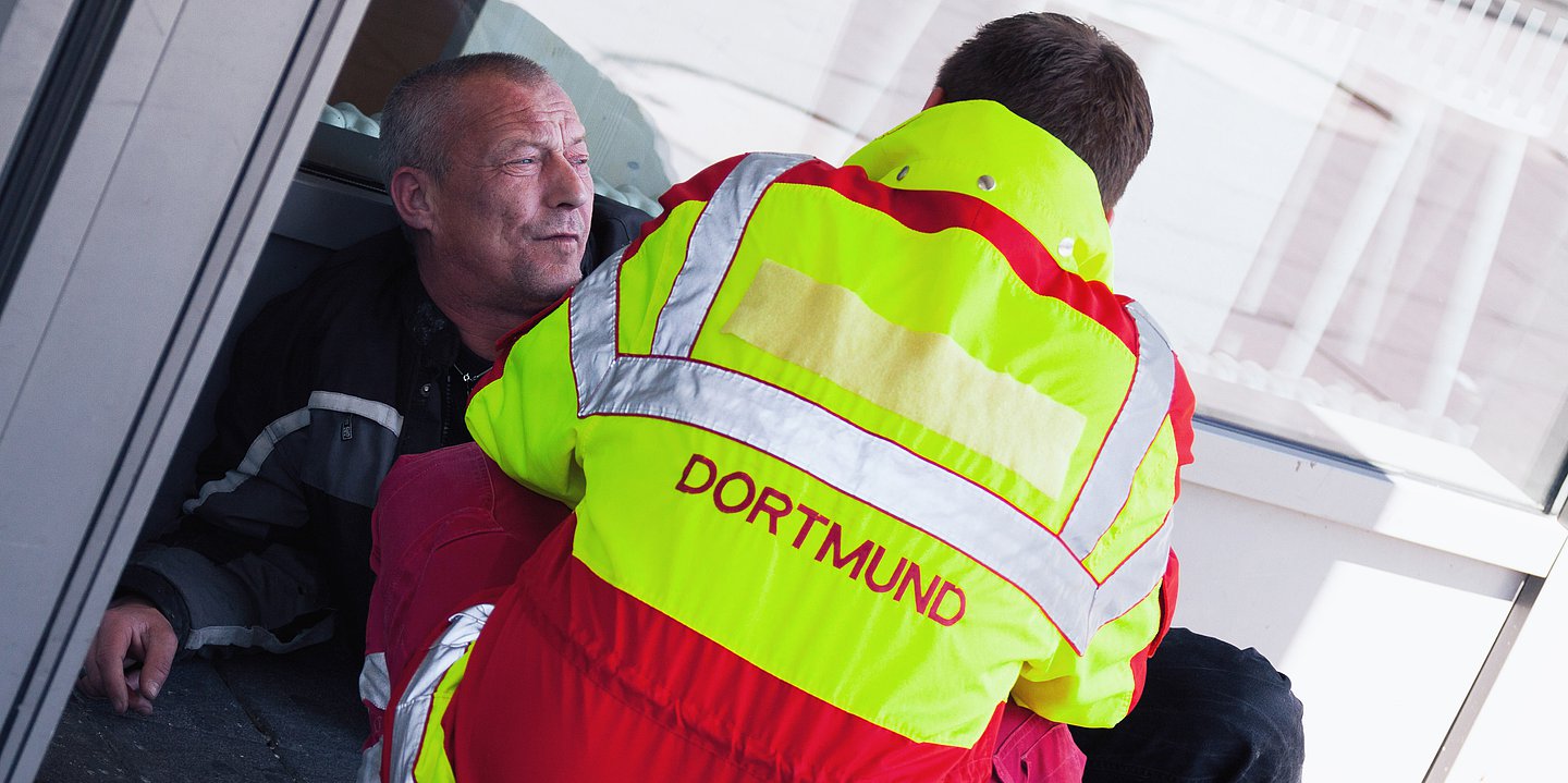 Foto: Eine obdachlose Person lehnt lehnt an einer Wand auf der Straße, davor kniet ein Rettungssanitäter. 