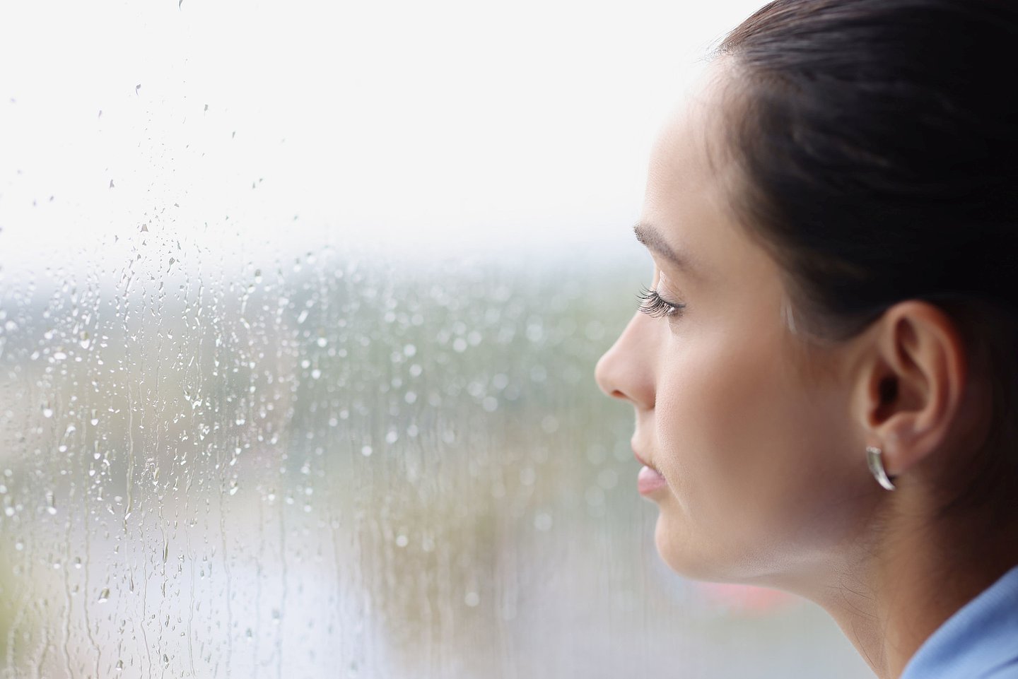 Das Foto zeigt eine junge Frau in Nahaufnahme, die traurig aus dem regennassen Fenster schaut.