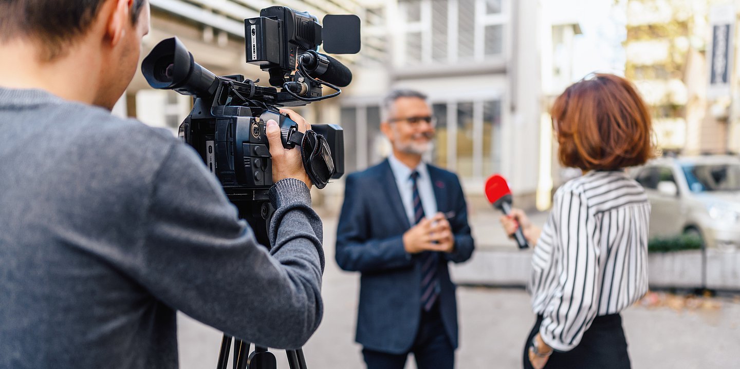 Das Bild zeigt rechts eine Journalistin mit rotbraunen, schulterlangen Haaren und weiß-schwarz längs gestreifter Bluse. Sie hat die linke Hand in ihrer Hosentasche. In der rechten hält sie ein Mikrofon mit rotem Windschutz und interviewt einen Mann mit graumeliertem Haar und Vollbart. Er trägt einen graublauen Anzug und ein hellblaues Hemd mit dunkler Krawatte. Im Vordergrund rechts steht ein Kameramann mit grauem Pullover, der die Szenerie mit dem Rücken zum Betrachter filmt.