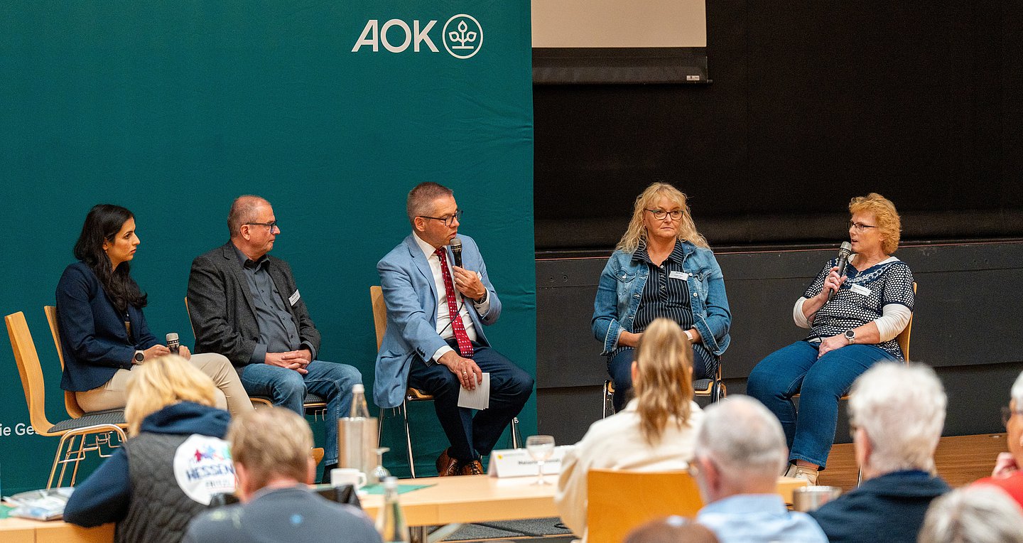 Das Bild zeigt eine Diskussionsrunde aus drei FRauen und einem Mann auf einem Podium plus einem Moderator in der Mitte vor einer dunkelgrünen Wand mit AOK-Logo.. Davor sitzen