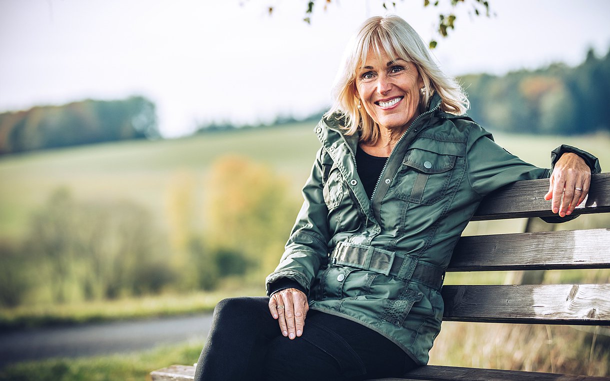 Das Bild zeigt eine lächelnde blonde Frau mit schulterlangen Haaren. Sie Sitzt unter einem Baum auf einer Bank, trägt einen schwarzen Rock, schwarze Strumpfhosen, ein schwarzes Top und eine grüne Outdoor-Jacke