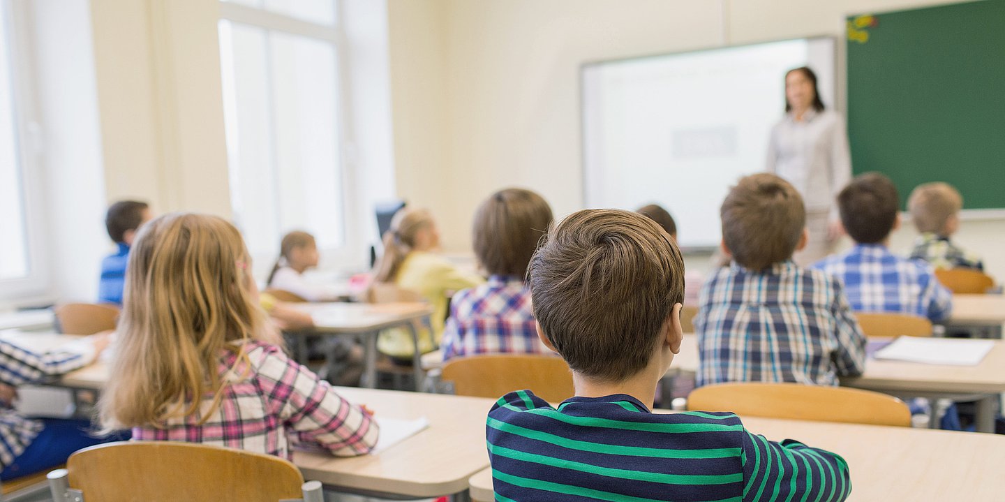 Foto: Eine Lehrerin steht vor einer Schulklasse, neben ihr ein Smartboard.