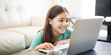 Medienkompetenz Junges Mädchen, das vor dem Sofa am Couchtisch sitzt und in einen Laptop grinst
