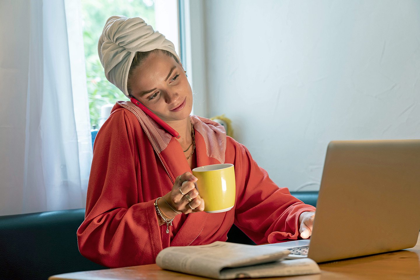 Foto: Eine junge Frau sitzt im Bademantel vor dem Laptop, mit Handy am Ohr und einer Kaffeetasse in der Hand. 