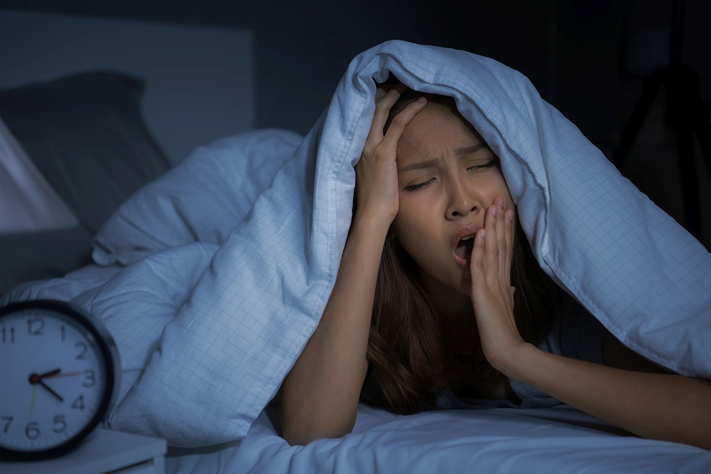 Foto zeigt eine Frau mitten in der Nacht, die gähnend die Bettdecke über den Kopf gezogen hat und nicht schlafen kann.