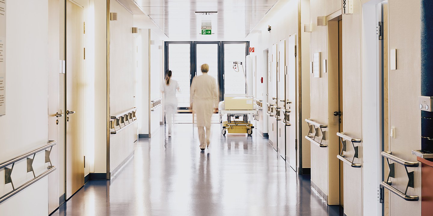 Foto eines hellen Krankenhausflurs druch den zwei Medizinerinnen laufen und in dem ein Bett steht