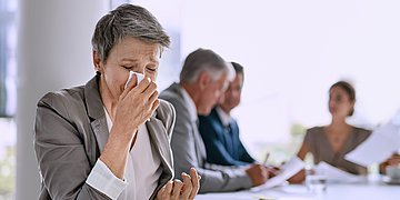 Präsentismus Eine Frau sitzt an einem Meetingtisch mit anderen Kolleginnen und Kollegen. Sie dreht sich weg und niest in ein Taschentuch.