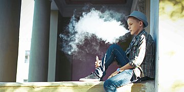 Foto: Ein Junge, etwa 11 Jahre alt, sitzt auf einer Mauer und raucht eine E-Zigarette.