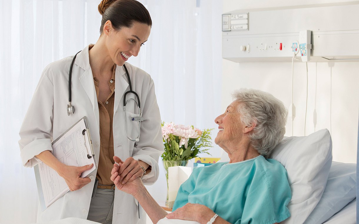 Foto einer Ärztin, die sich mit einer Frau im Krankenbett unterhält. Beide wirken zuversichtlich.