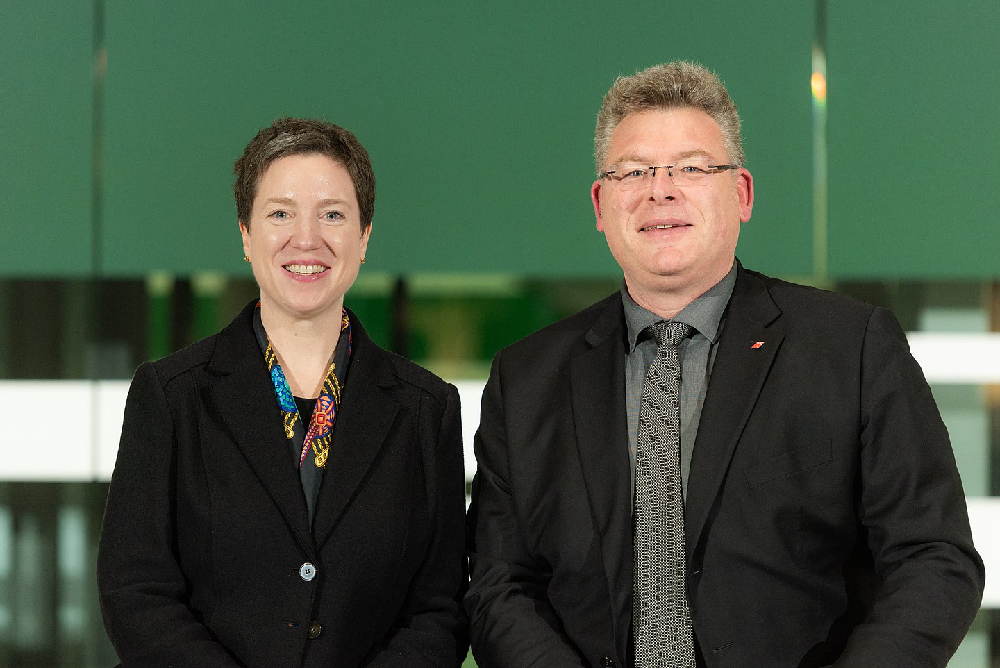 Foto zeigt Dr. Susanne Wagenmann links mit brünettem Kurzhaarschnitt und bunter Bluse. Rechts im Bild Knut Lambertin mit Brille und graumelierten kurzen Haaren. Knut Lambertin trägt ein graues Hemd mit grauer Krawatte. Beide tragen ein schwarzes Sakko.