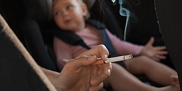 Foto eines Kleinkindes im Auto, davor eine Hand mit einer brennenden Zigarette