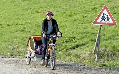 Foto: Eine Frau macht einen Fahrradausflug mit ihren Kindern, die in einem Fahrradanhänger sind. Alle drei Personen tragen einen Schutzhelm.