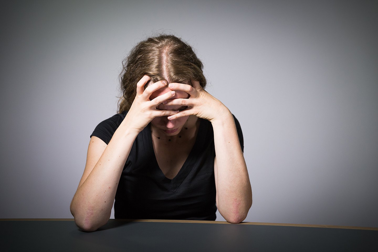 Das Bild zeigt eine junge, blonde Frau mit schwarzem Top an einem Tisch vor einer graus-schattierten Wand sitzend. Sie stützt ihren Kopf auf den Armen und verbirgt dabei ihr Gesicht