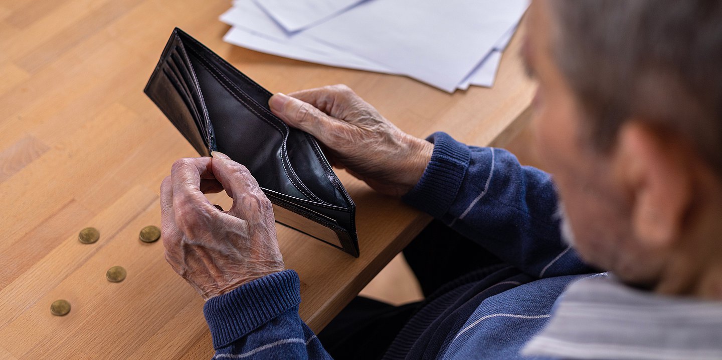 Foto: Ein älterer Mann schaut in seinen leeren Geldbeutel, daneben liegen einige Münzen auf einem Tisch.