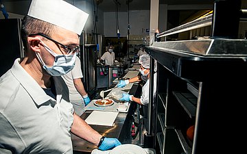 Klinikkantine Havelhöhe Foto von Küchenmitarbeitenden, die mit Maske Tabletts in einer Klinikkantine befüllen