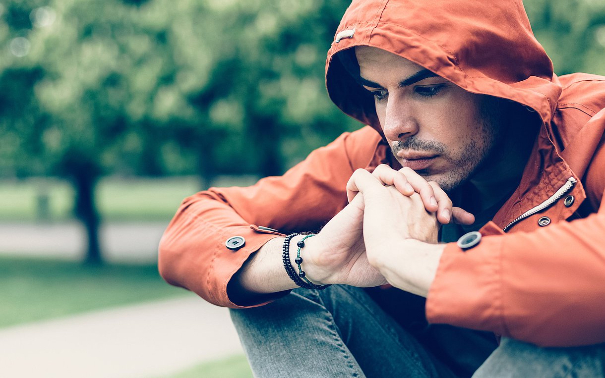 Foto eines Mannes in einer orangenen Kapuzenjacke, der verzweifelt in einem Park sitzt