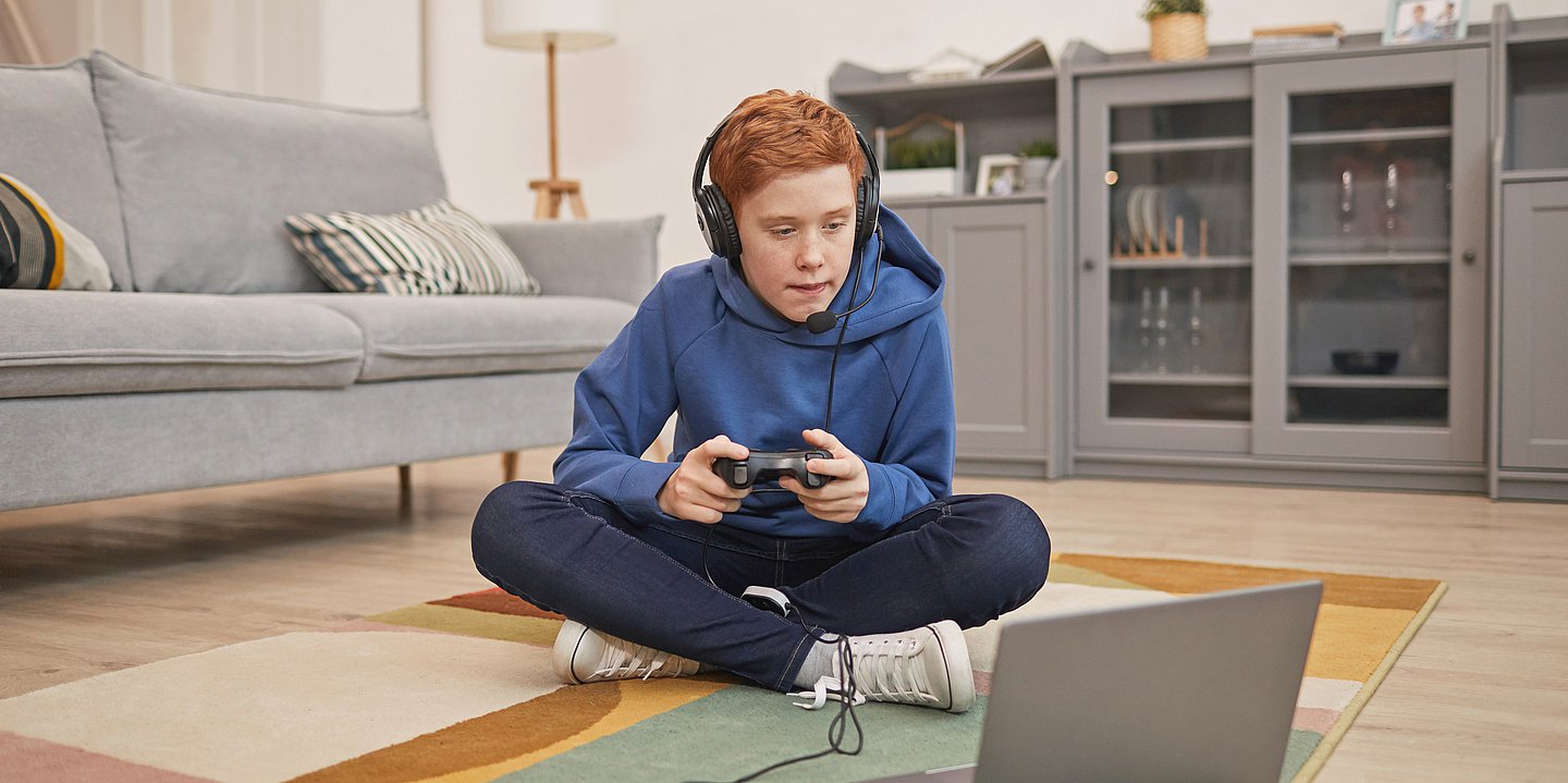 Foto: Ein Junge sitzt mit einem Controller und Kopfhörern im Schneidersitz vor einem Laptop.