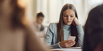 Eine Schülerin schaut während des Unterrichts gelangweilt auf ein Smartphone.