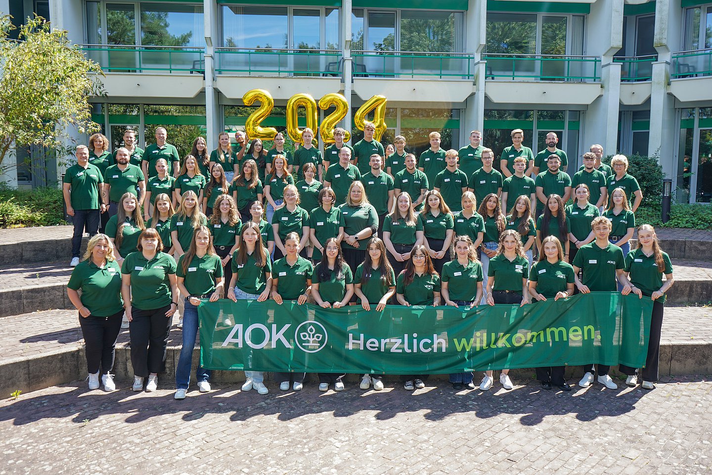 Das Gruppenbild zeigt den Ausbildungsjahrgang 2024. Die Azubis in vier aufsteigenden Reihen tragen alle das grüne AOK-Poloshirt. Die vorderste Reihe hält ein Banner mit dem AOK-Bäumchen und der Aufschrift "Herzlich Willkommen". Oberhalb der Gruppe hängen goldene Luftballons, die die Jahreszahl 2024 bilden.