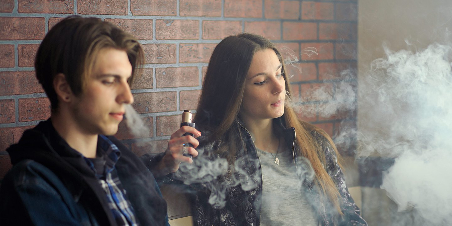 Foto: Ein junger Mann und eine junge Frau sitzen nebeneinander und rauchen E-Zigarette.