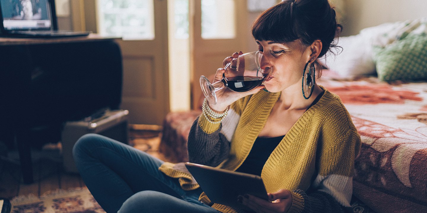 Foto: Eine Frau schaut auf ihr Tablet und trinkt dabei Rotwein.