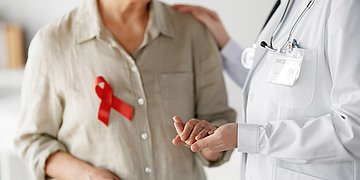 HIV Foto einer älteren Dame mit HIV-Schleife, die eine Medizinerin an der Hand hält. Die Gesichter sind nicht zu sehen.