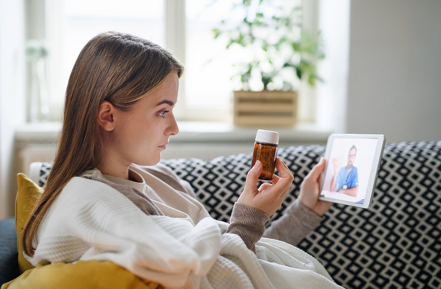 Junge Frau sitzt zu Hause auf dem Sofa und hat eine Videosprechstunde mit ihrem Arzt. Gerade hält sie ein kleines braunes Medizin-Fläschchen für den Arzt in die Kamera.