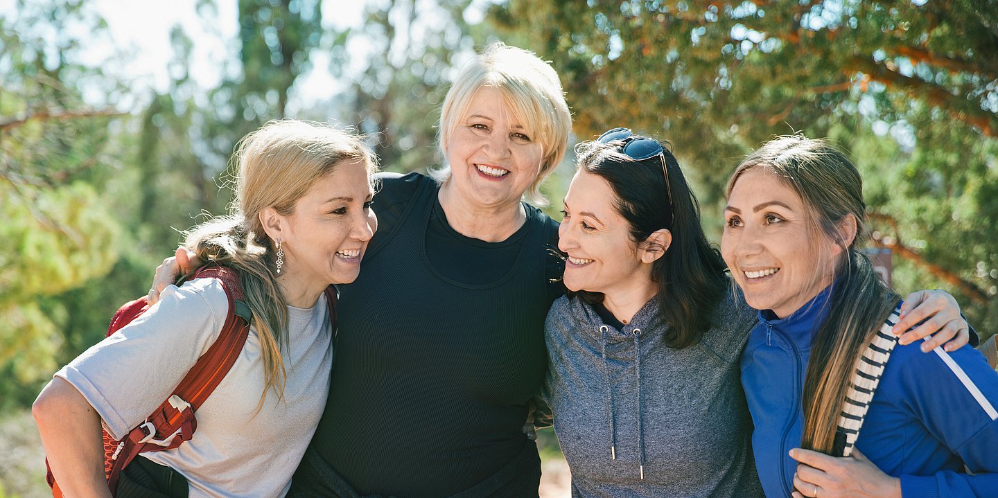 Vier Frauen stehen lachend im Park und legen die Arme umeinander.