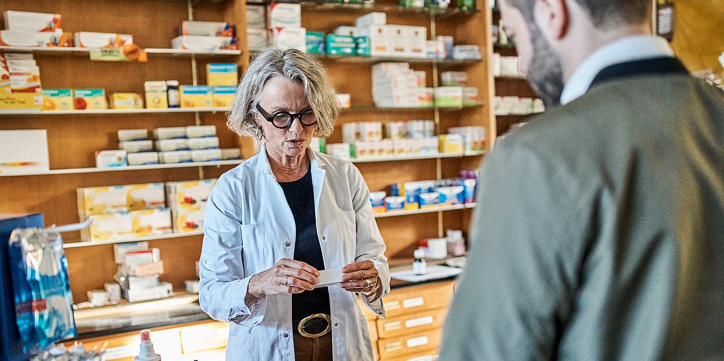 Foto einer Apothekerin, die vor einem Regal mit Medikamenten steht und einen Kunden bedient