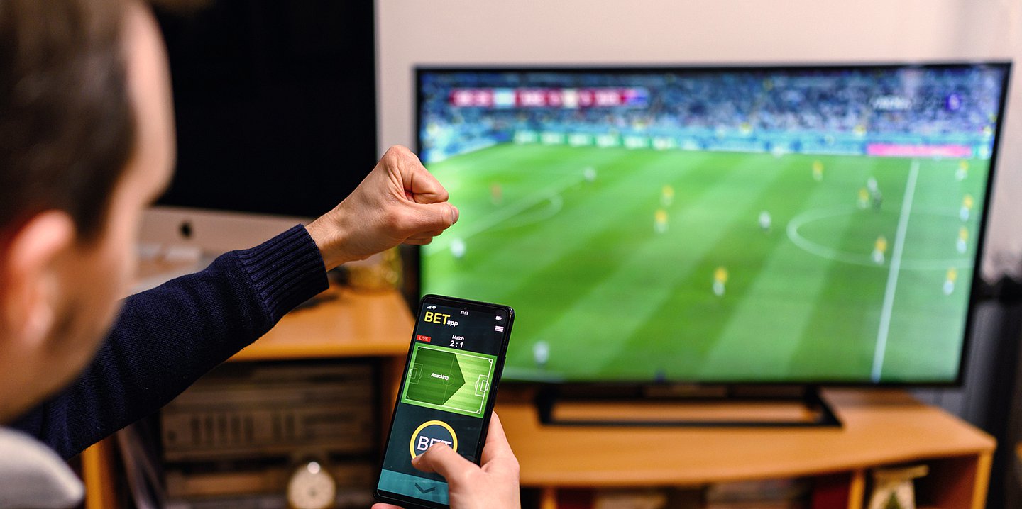 Foto: Ein Mann sitzt mit seinem Smartphone, auf dem eine App für Sportwetten zu sehen ist, vor dem TV-Bildschirm, auf dem ein Fußballspiel läuft.