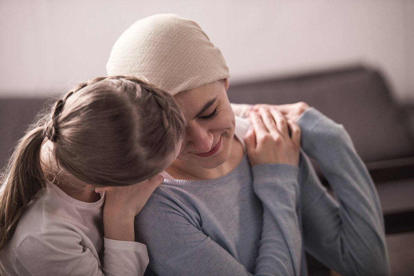 Foto zeigt eine kranke junge Frau auf einem Sofa sitzend, die sich an eine sich ihr zuwendende Frau anlehnt, die sie an den Schultern hält.