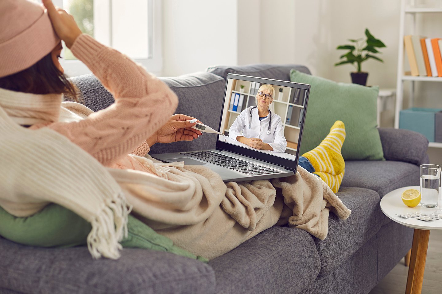 Foto zeigt eine kranke Frau von hinten, die auf einem Sofa liegend sitzt. Sie trägt eine Wollmütze auf dem Kopf, einen Schal um den Hals und ist in eine Wolldecke gewickelt. Auf dem Schoß hat sie einen Laptop, der Bildschirm zeigt eine Ärztin im Gespräch mit der Frau, die in der linken Hand ein Fieberthermometer hält und sich mit der rechten Hand an die Stirn fasst.