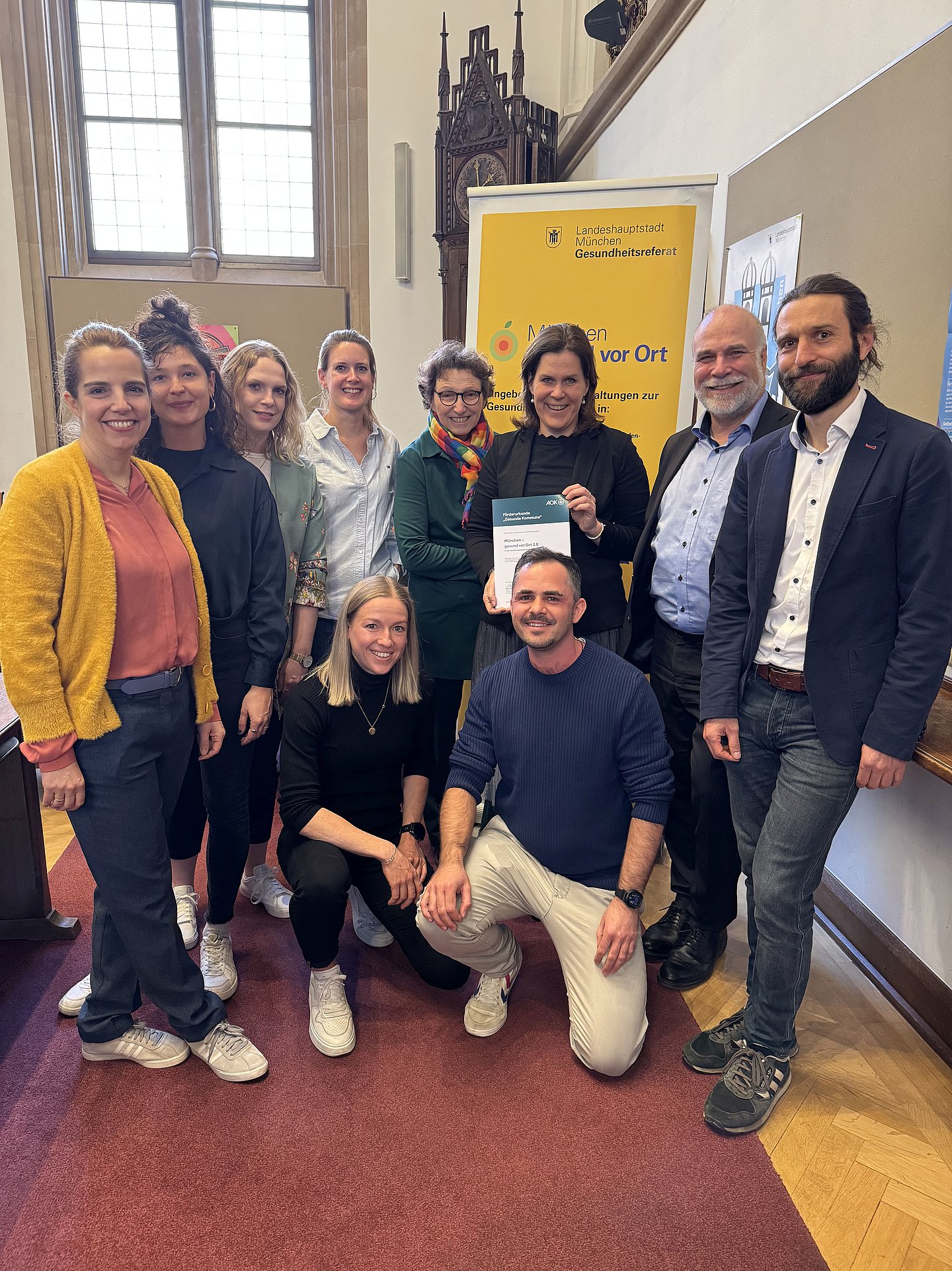 Im Foto stehend von links nach rechts in der oberen Reihe: Christina Schub mit Greta Wirth und Bernadette Hülsmann (alle vom Projekt "München – gesund vor Ort"), Dr. Verena Lindacher (Abteilungsleitung Stadtteilgesundheit), Beatrix Zurek (Gesundheitsreferentin), Verena Dietl (Bürgermeisterin), Martin Felber (Direktor der Direktion München, AOK Bayern) und Andreas Nagel (Referent Lebenswelten, AOK Bayern) sowie in der unteren Reihe Lena Donhauser und Sven von Braumüller (beide: Projekt "München – gesund vor Ort")